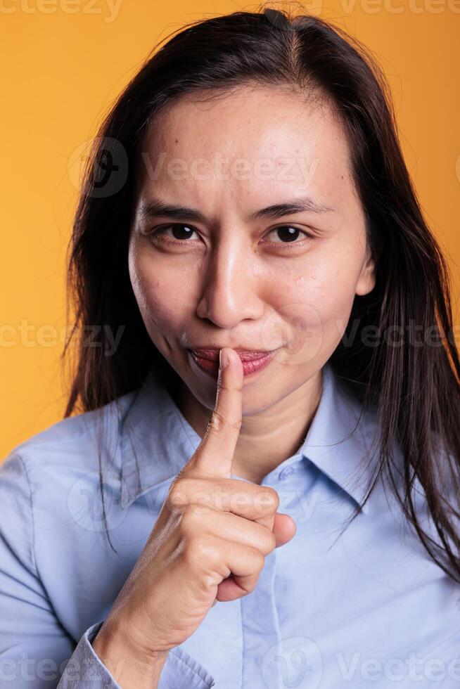 Cheerful serious filipino woman making silence gesture by putting forefinger over lips posing in studio over yellow background. Young adult doing shh symbol, expressing confidentiality photo