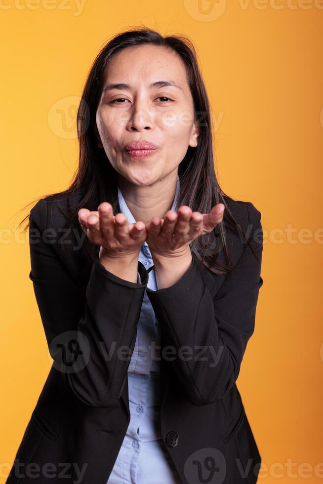 Filipino woman blowing air kissed during studio shot, posing over yellow background. Confident positive young adult expressing love, looking at the camera with a satisfied smile. Romantic gesture photo