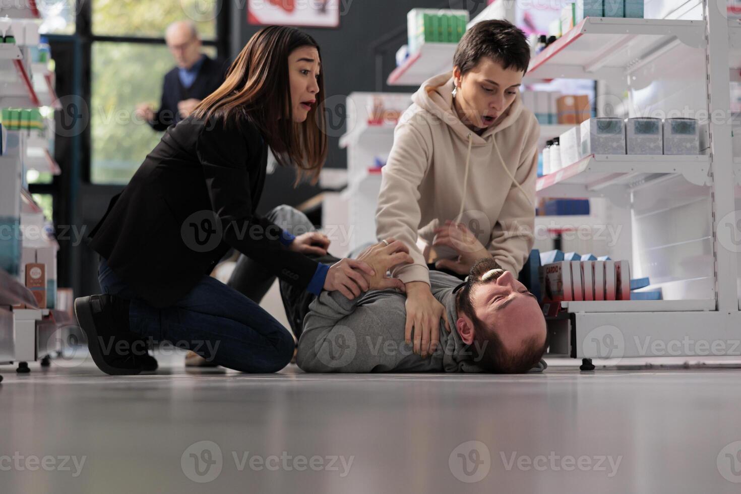 Frightened women rescuing man suffering from epilepsy attack, holding unconscious person and waiting for help in drugstore. Pharmacy asian and caucasian clients noticing epileptic seizure accident photo