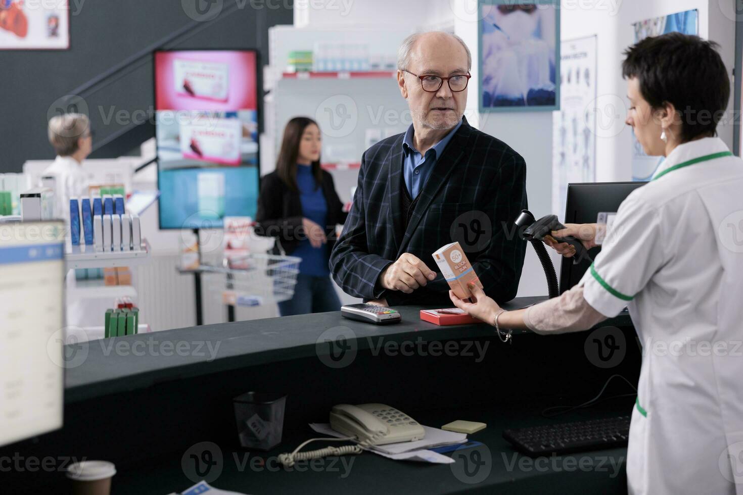 Elderly man giving pharmacy cashier medical product for scanning at checkout and discussing sunscreen with pharmaceutical worker. Customer visiting drugstore to buy sun protection lotion and vitamin photo