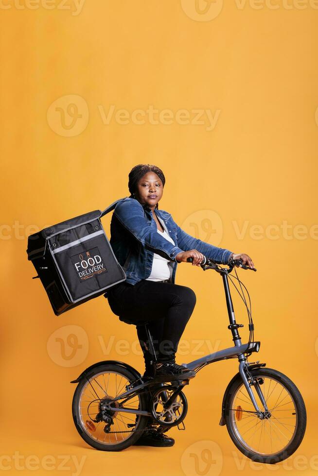 Restaurant worker riding bike while carrying takeaway thermal backpack ready to deliver order to customer during lunch time, standing over yellow background. Take out concept and transportation photo