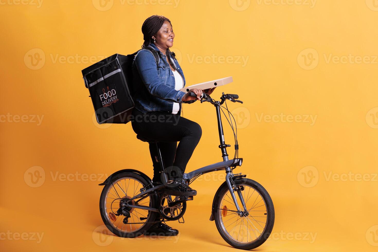 African american pizzeria courier carrying pizza carton box delivering takeaway order to client during lunch time, using bike as transportation in studio. Take out food service and concept photo