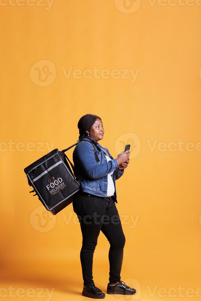 African american deliverywoman holding phone looking at customer adreess on fast food app before start deliver takeout order. Pizzeria courier carrying takeaway thermal backpack in studio photo