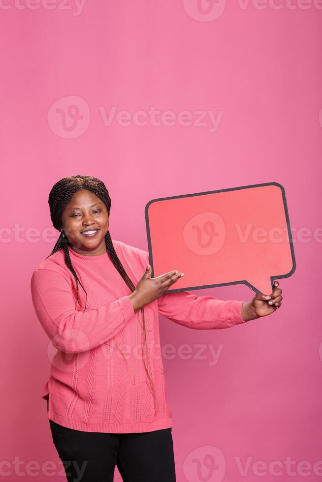 Smiling african american confident woman holding red cardboard speech bubble standing in studio on pink background. Joyful young adult holding blank template announcement placard. photo