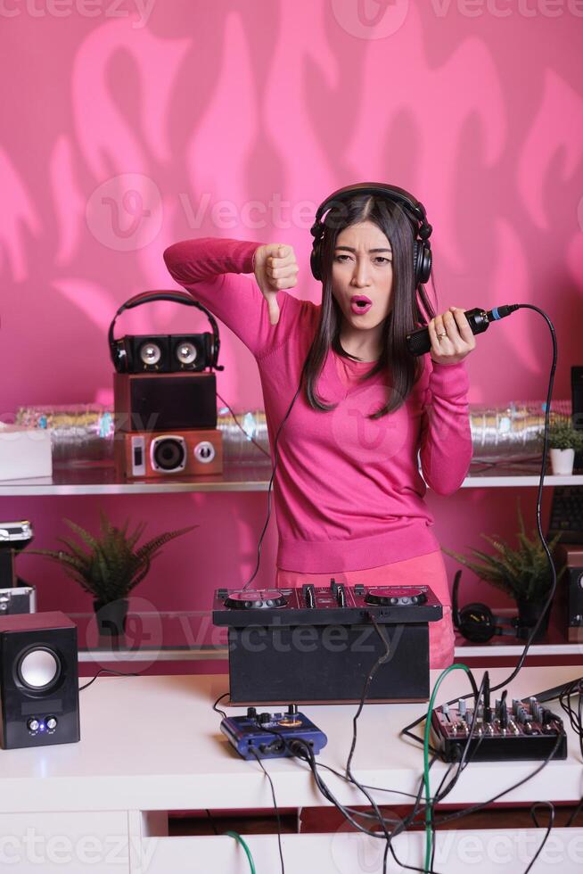 Asian perfomer showing disagreement sign in front of camera while mixing electronic sounds with techno using professional mixer console. Artist doing performance at nightclub with audio equipment photo