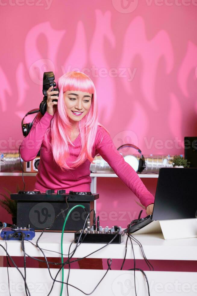 Asian artist having fun while performing eletronic music in night club playing song using professional turntables. Woman standing at dj table dancing while celebrating techno album with fans photo