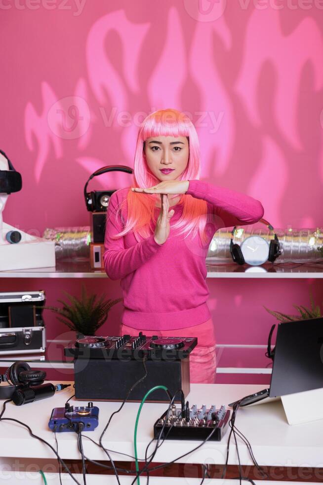 Asian musician standing at dj table doing time out gesture with palm while performing techno song using professional mixer console in club at night. Woman artist having serious expression, break sign photo