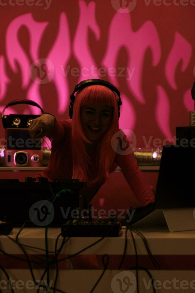 Artist with pink hair wearing headset while playing techno song at professional mixer console, enjoying night life in studio with pink background. Asian dj performing electronic music in club photo