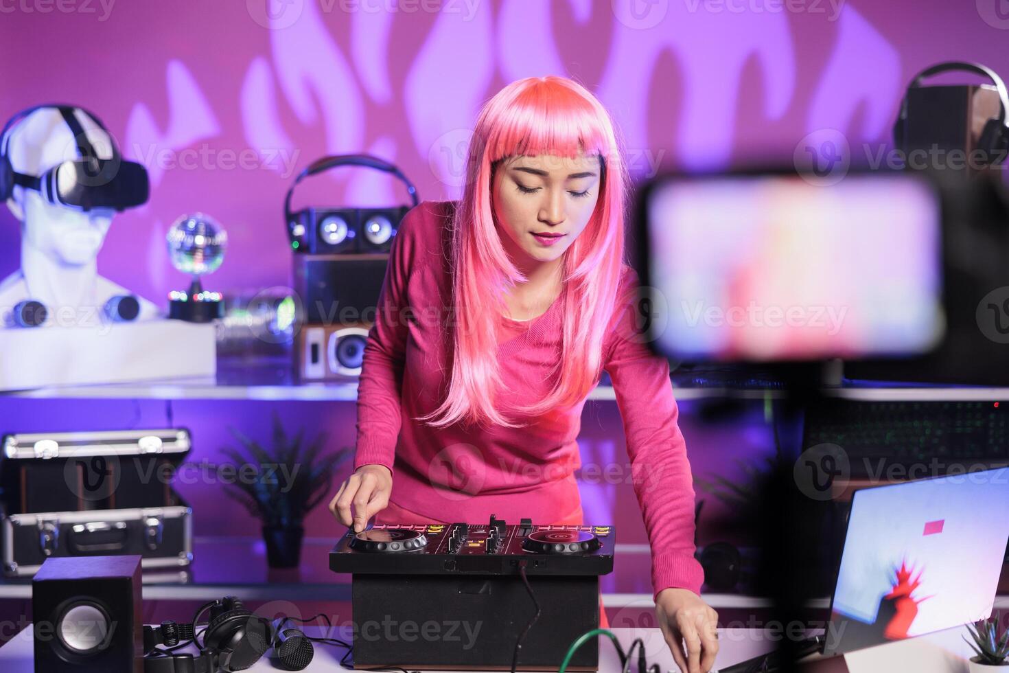 Smiling woman mixing techno music at professional mixer console having fun while performing song at night in club. Artist with pink hair recording performace with camera posting online for subscribers photo