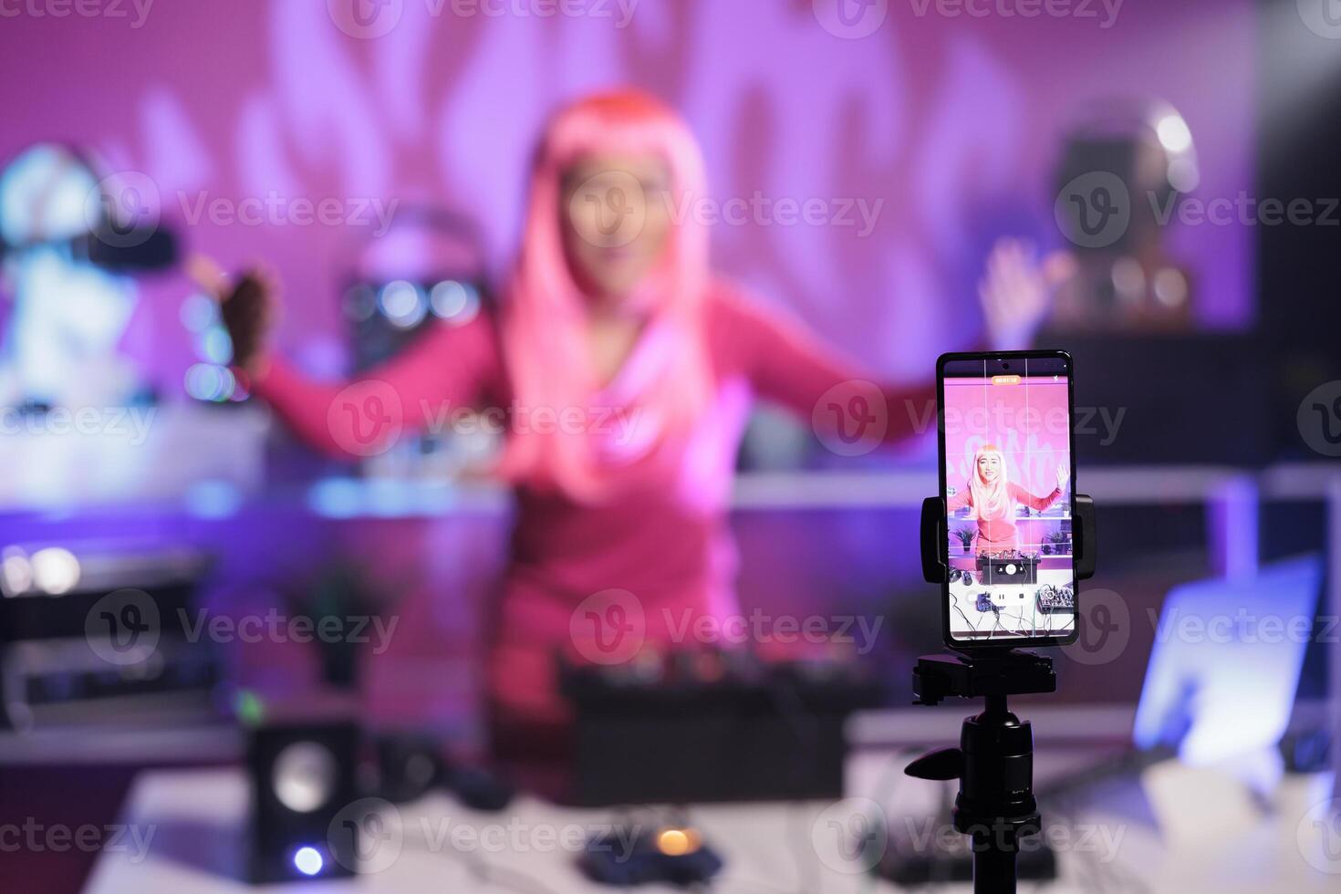 Woman with pink hair standing at dj table mixing song with turntables in front of camera while recording video with phone. Smiling artist doing performance at nightclub with audio equipment photo