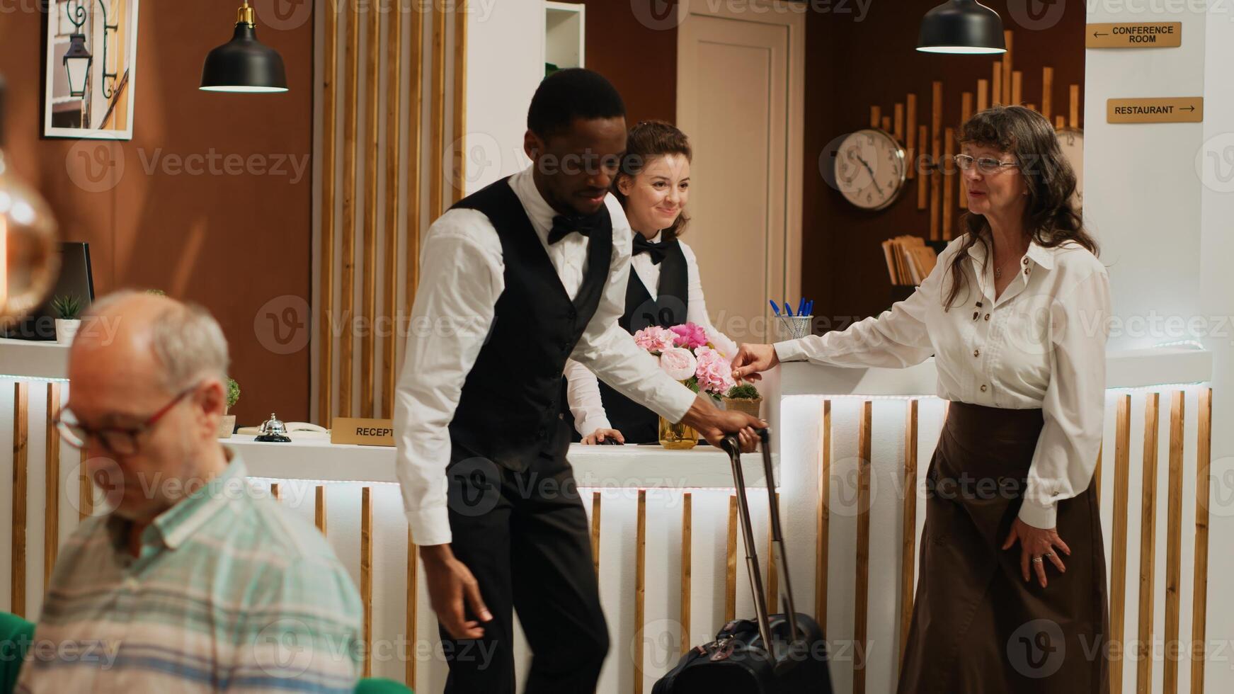 Senior guest arriving at hotel with room reservation, bellboy offering luxury concierge services and carrying luggage for client. Receptionist doing check in process with traveller at front desk. photo