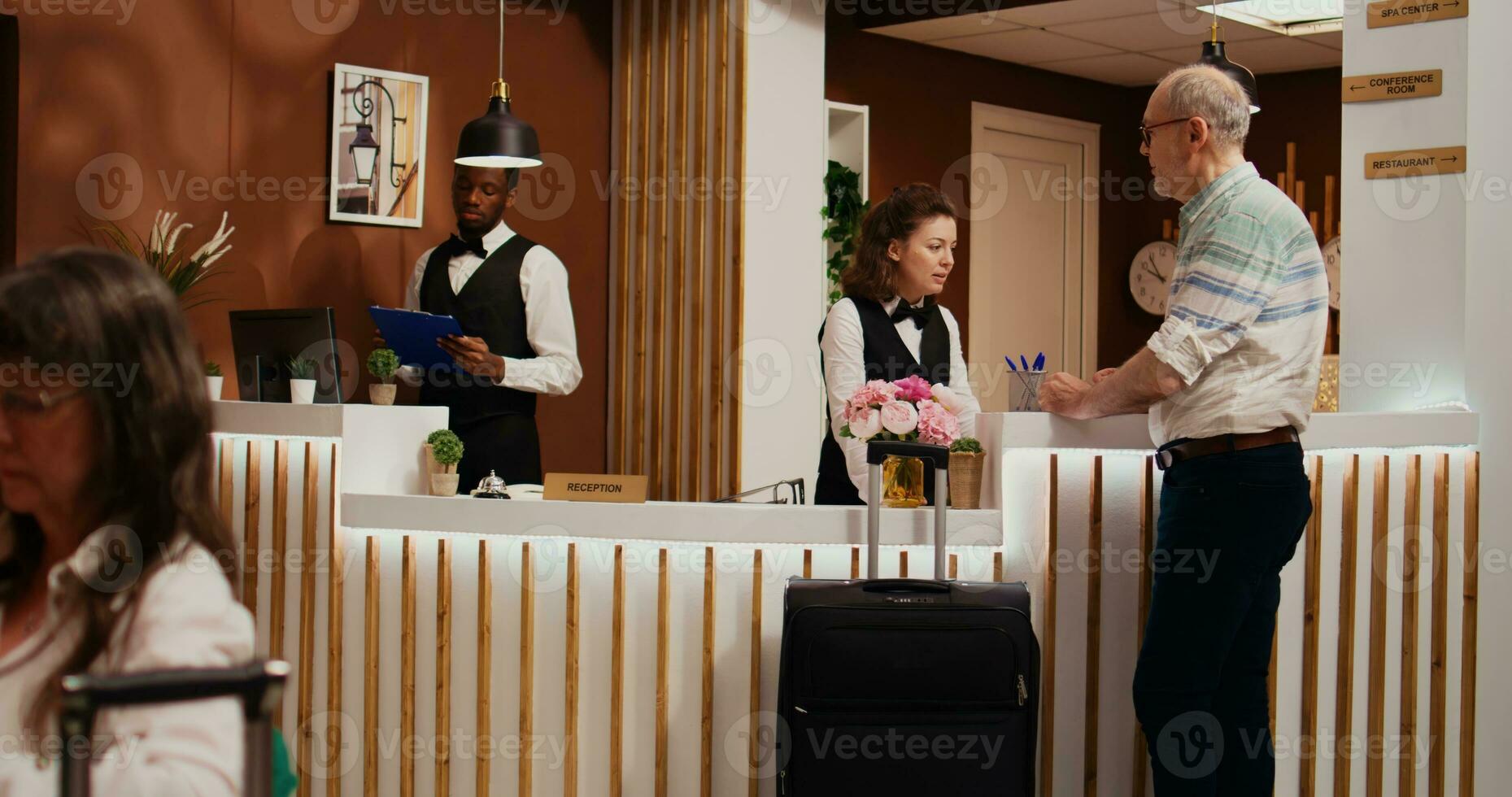 Senior man entering hotel lobby with luggage, asking for concierge services to check in. Receptionist woman ensuring relaxing stay for old tourist at all inclusive holiday resort, tourism industry. photo