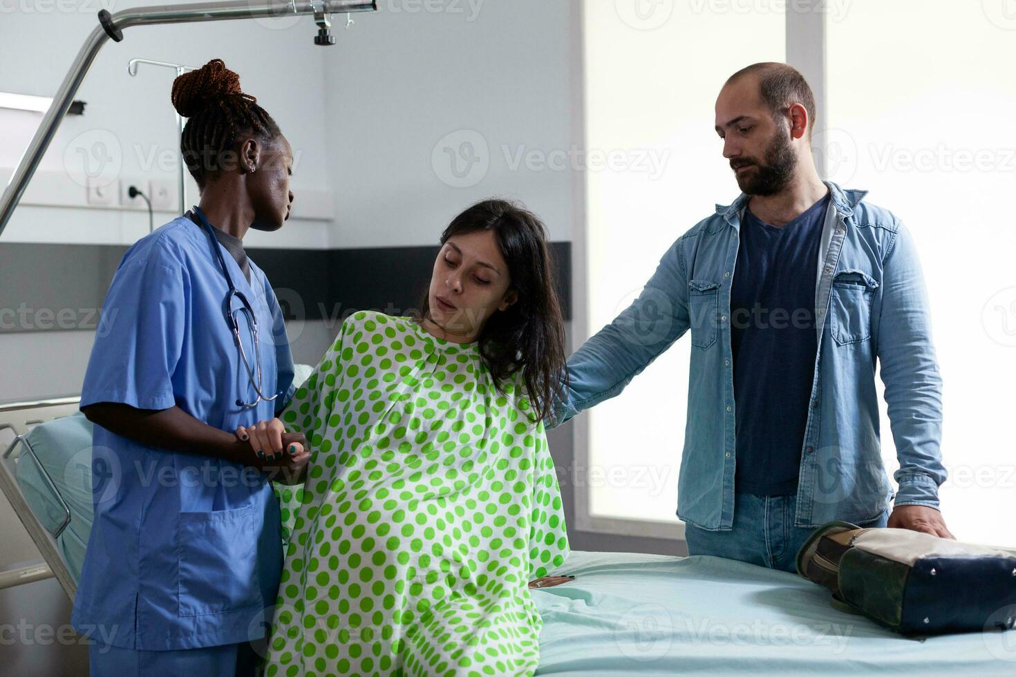 africano americano asistente Ayudar embarazada mujer a obtener fuera de cama en hospital pabellón, preparando paciente para cesariano cirugía. futuro padres esperando niño en maternidad clínica foto