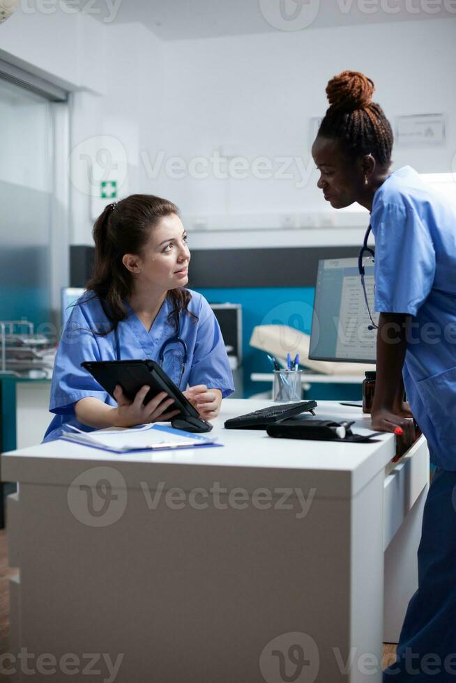 Nurse colleagues in happy relaxed hospital workspace working on tablet in modern professional medical office. Diverse healthcare specialists coworkers in modern clinic using technology photo