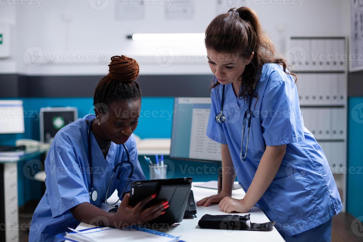 Nurse showing medical data to young collegue on a digital tablet screen in modern professional clinic office. Diverse clean modern hospital workplace equipped with technology photo