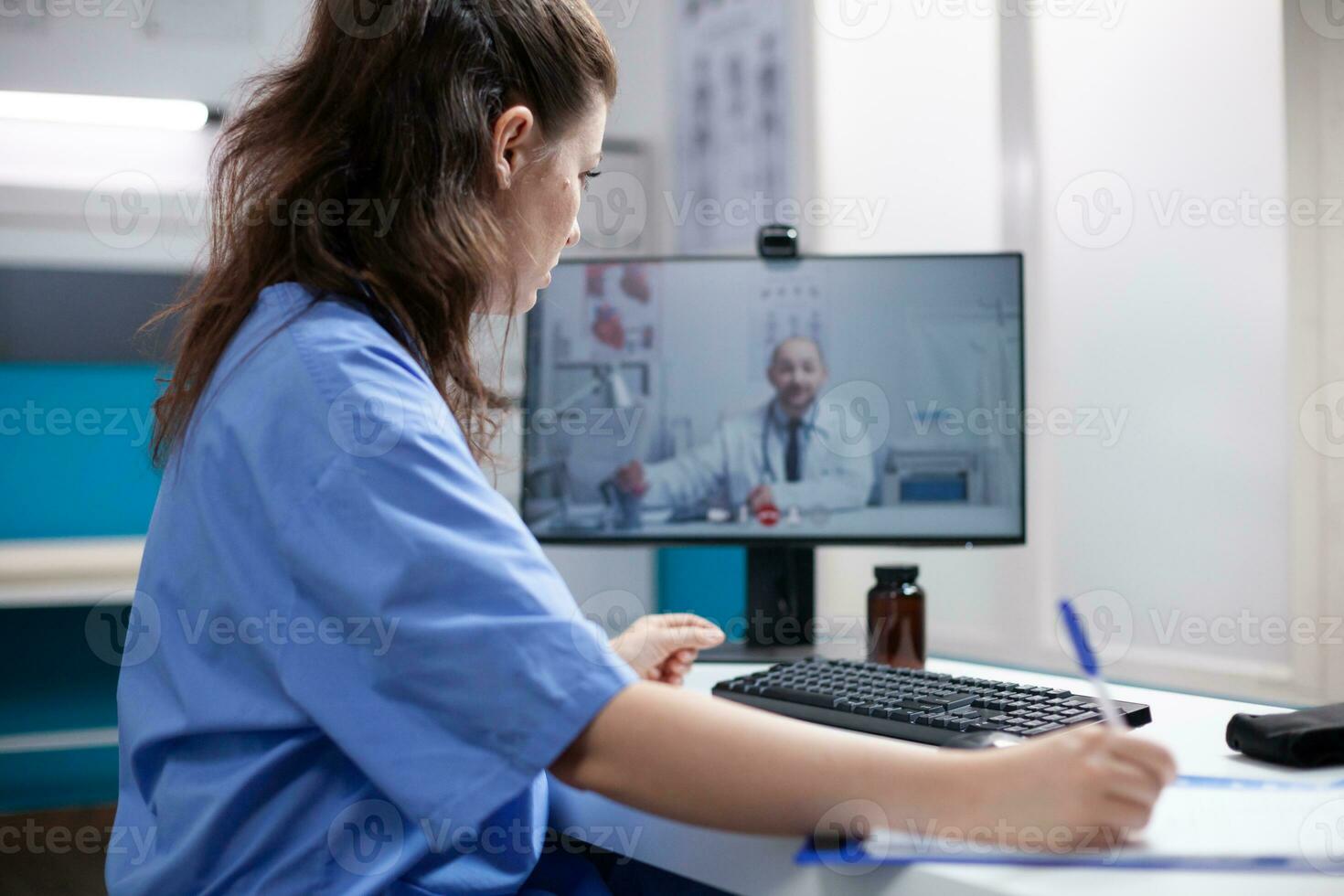 Nurse in videocall taking notes from doctor colleague in professional medical office. Woman writing down information received from general practitioner over telemedicine videoconference photo