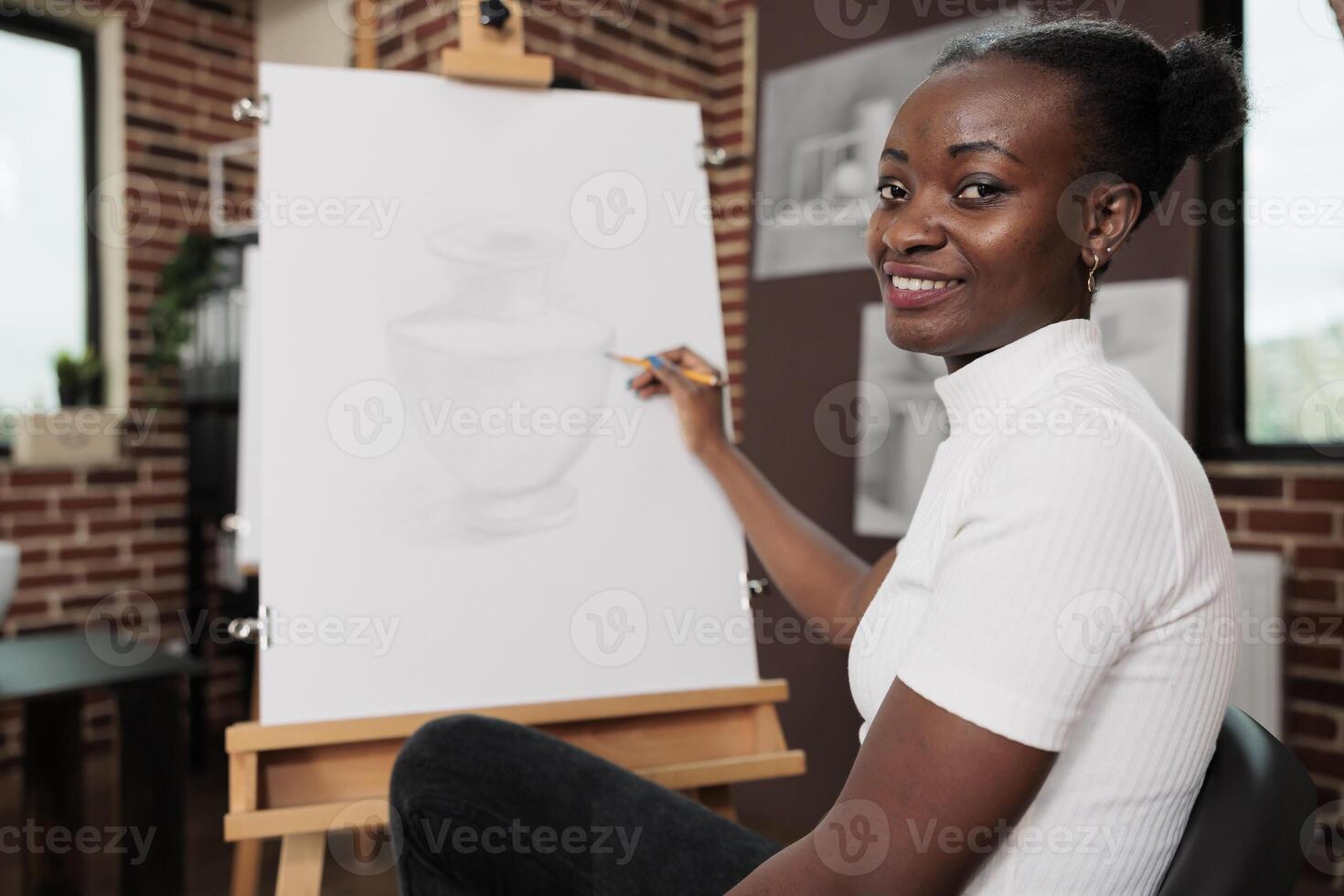 Happy inspired young African American woman creating unique piece of art during creative art workshop, sitting at easel and smiling at camera, enjoying drawing lesson, developing her own creativity photo