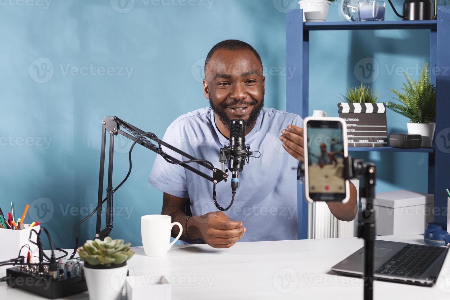 Smiling blogger streaming and looking at smartphone camera while creating digital content for online blog. Internet streamer talking and using mobile phone on tripod in studio photo