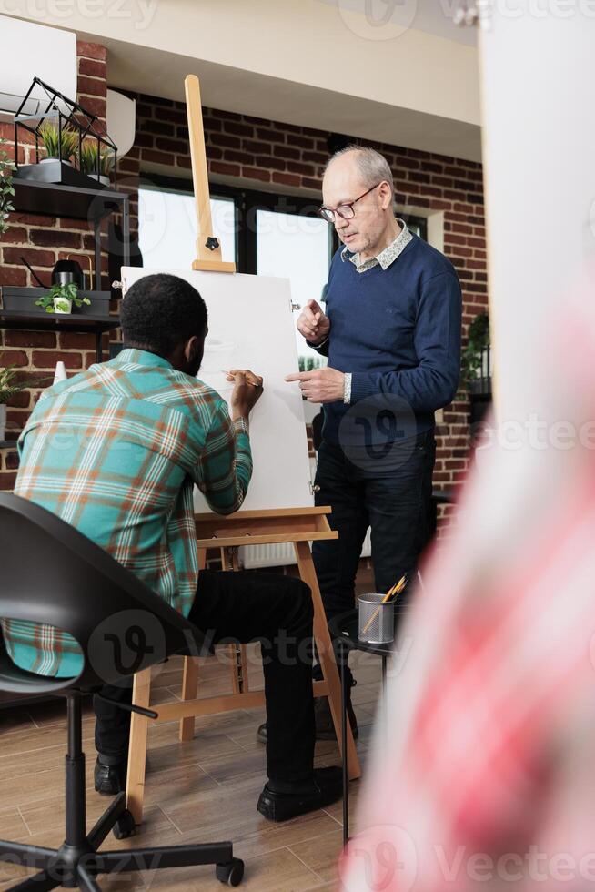 Senior man art teacher standing near easel working with African American male student during drawing lesson, experienced instructor teaching people to sketch. Artistic creative activities for adults photo