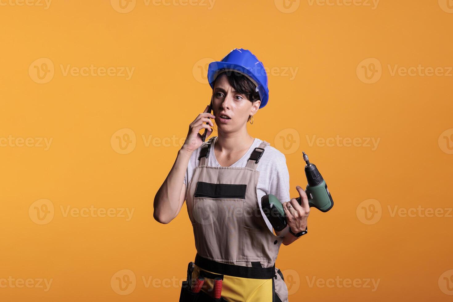Surprised amazed builder talking on phone call and holding drilling gun, using smartphone in studio. Female contractor answering mobile phone for remote chat before renovating project. photo
