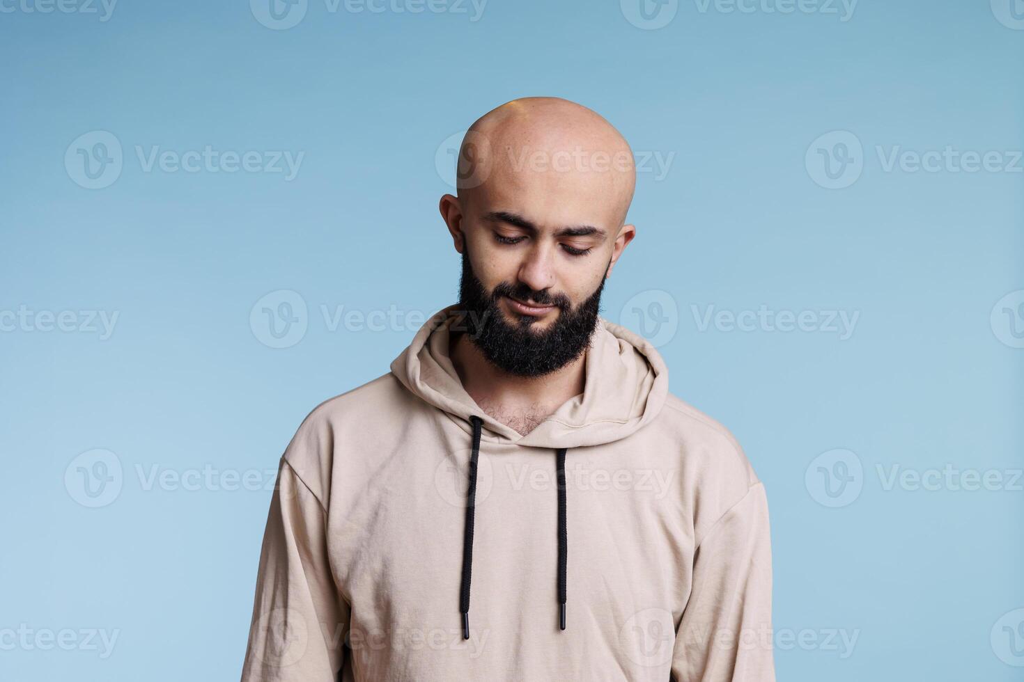 Arab man with neutral expression looking down and tilting head. Pensive young person wearing casual beige hoodie clothes thinking and standing in casual pose on blue background photo