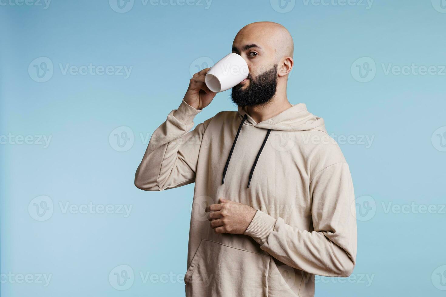 Young arab man drinking coffee from white cup and looking at camera. Arabian bald bearded person wearing casual clothes sipping tea, enjoying hot beverage from mug studio portrait photo