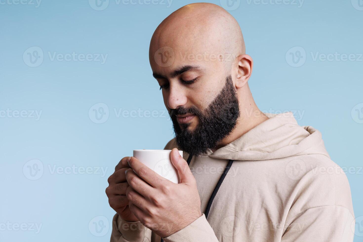 Thoughtful arab man holding cup and looking at coffee with pensive facial expression. Young adult handsome bald bearded person enjoying tea, drinking hot beverage from white mug photo