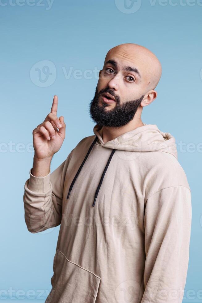 inspirado árabe hombre teniendo idea y levantamiento índice dedo arriba mientras mirando a cámara. joven calvo barbado persona con emocionado facial expresión señalando hacia arriba estudio retrato foto