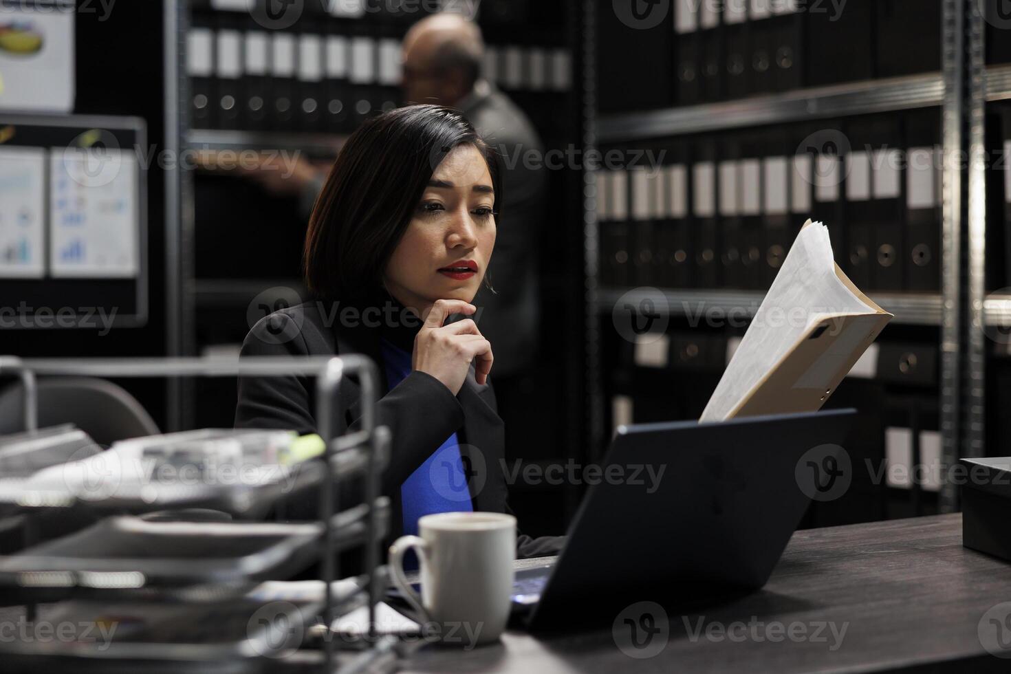 Bureaucratic administration employee checking accountancy budget plan data paperwork on laptop. Asian businesswoman executive in repository filled with document folders and graphical chart reports photo
