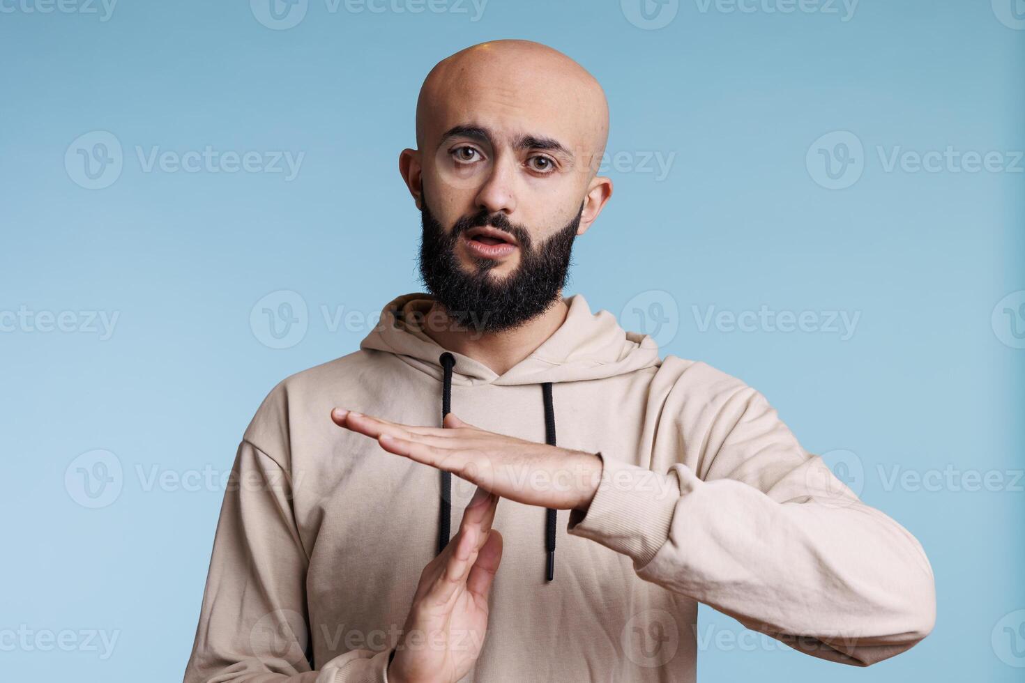 Arab man making time out gesture with hands while looking at camera. Young bald bearded person pausing conversation, showing interruption signal with arms studio portrait photo