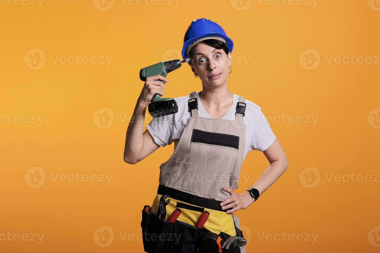 Female builder in hard hat holds electric drill against her head, going crazy from so much work. Professional woman construction worker wearing coveralls and tool belt in studio shot. photo