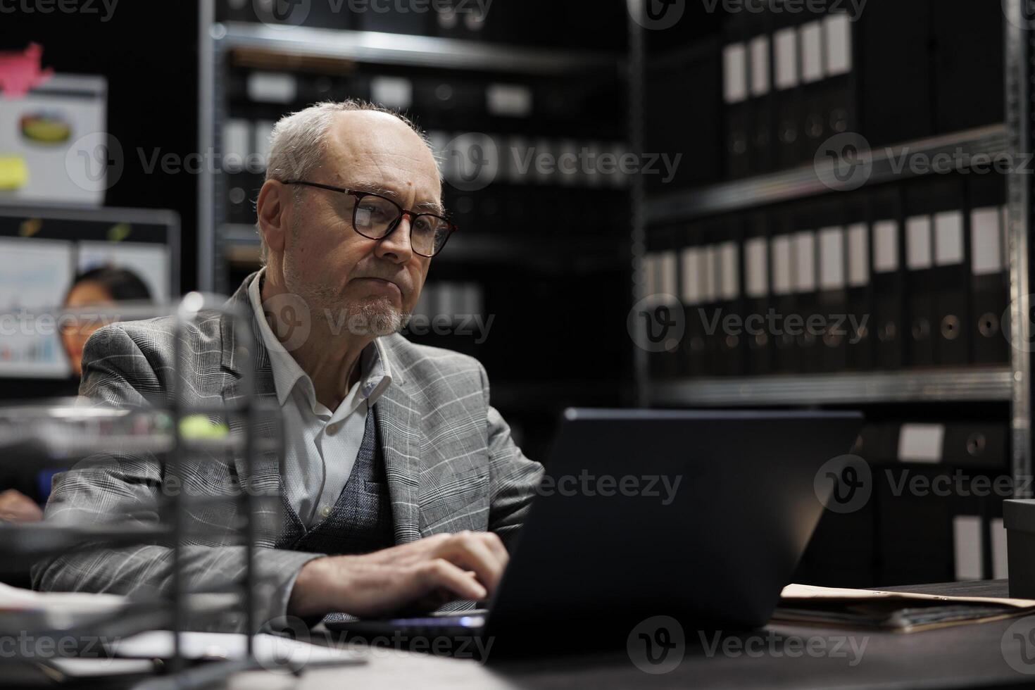 Senior private detective checking confidential criminology evidence files on laptop. Investigator in agency file storage office surrounded by folders on cabinet shelves photo