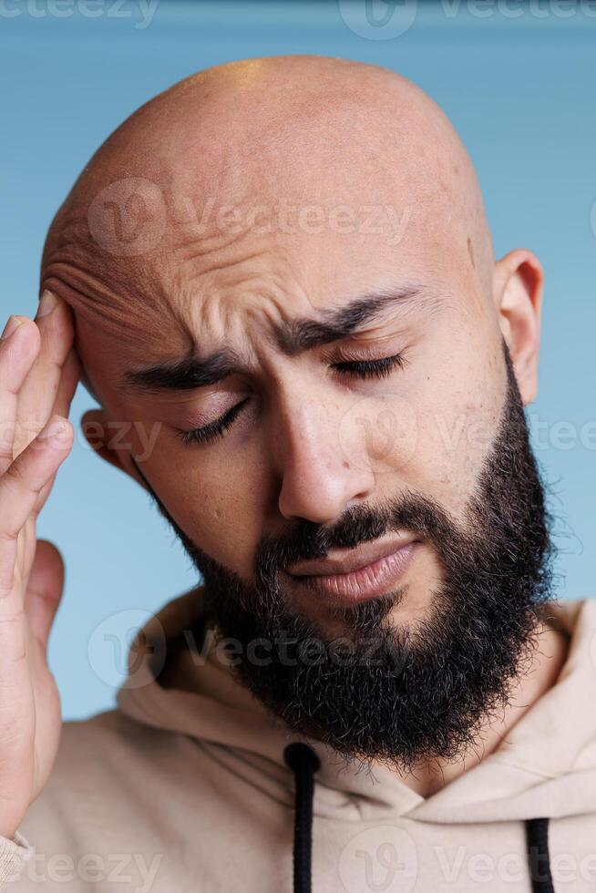 Arab man with closed eyes having migraine and rubbing head in pain. Young adult bald bearded person suffering from headache and holding temple while posing on blue background photo