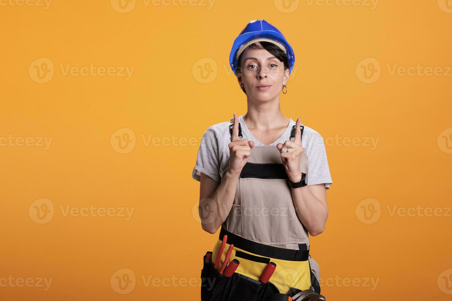 Portrait of woman builder pointing upwards with index fingers, showing above and indicating direction up. Posing in studio over yellow background and wearing hardhat with overalls. photo