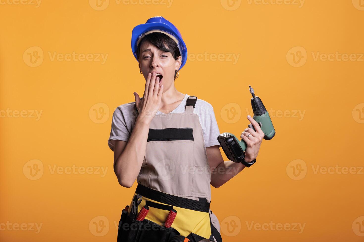 Sleepy female construction worker yawning and being tired while she works on renovating or building. Falling asleep and feeling exhausted, holding power drilling nail gun in studio. photo