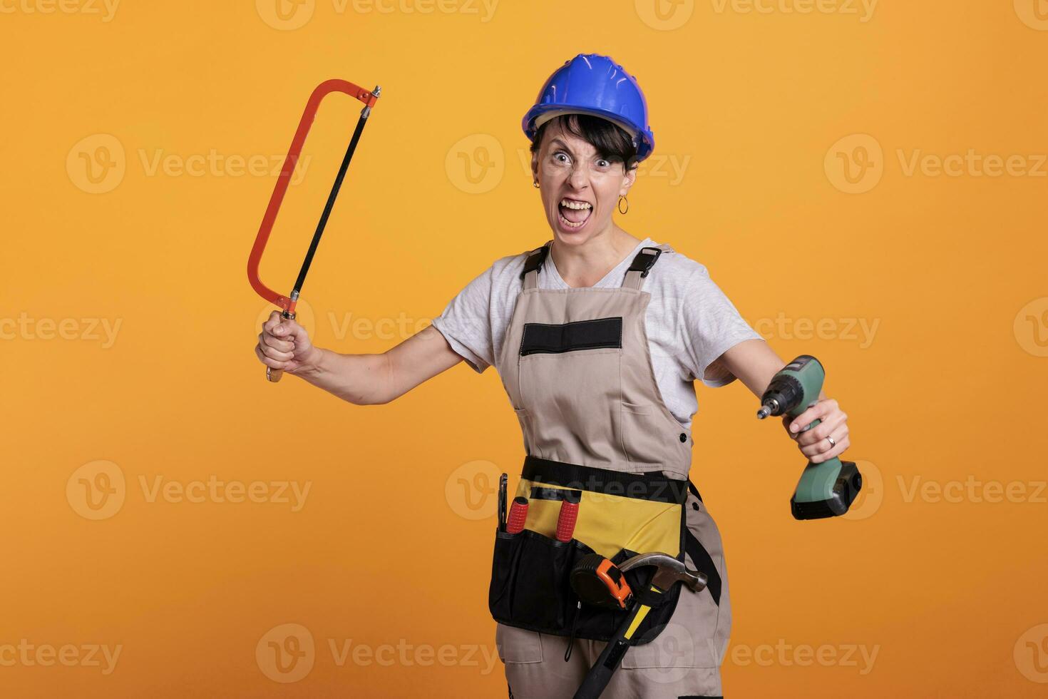 Angry violent woman builder with chainsaw and power drill acting crazy in studio, posing with building tools. Mad female worker being frustrated with manual saw and drilling gun. photo