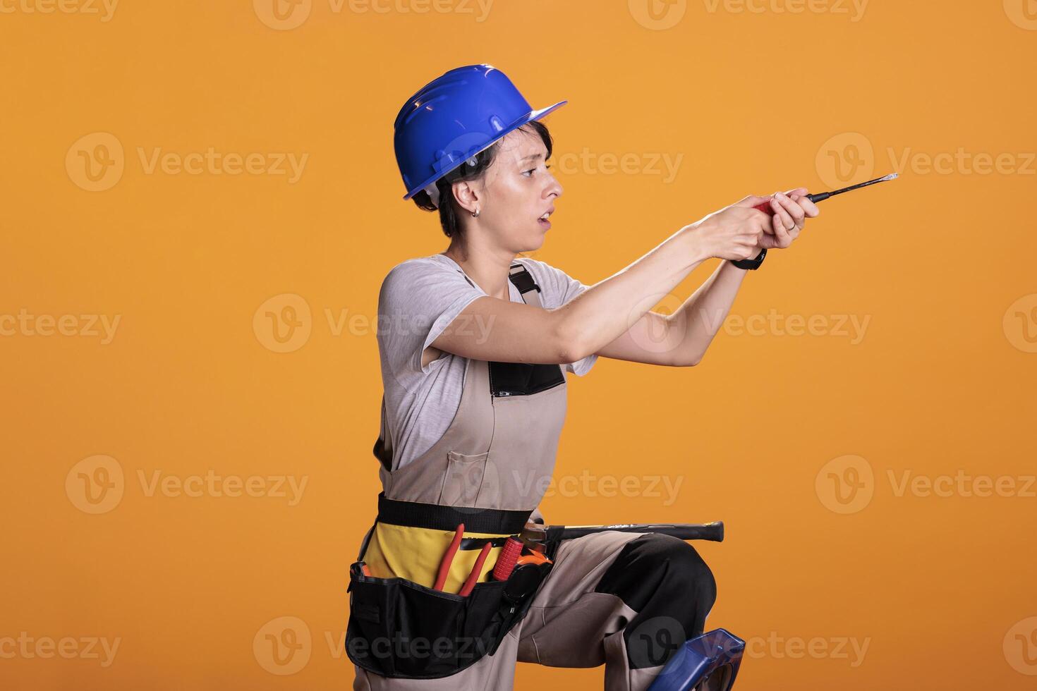 Construction worker on ladder repairing wall with screwdriver, posing in studio. Woman professional building contractor in uniform and hardhat using drill tools to screw nails. photo