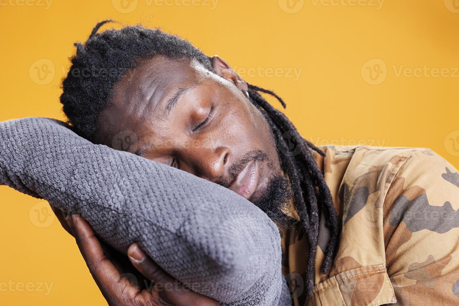 Exhausted young adult putting head on comfortable pillow to sleep and take nap. African american man trying to get rest after passing bedtime, sleeping in studio over yellow background photo
