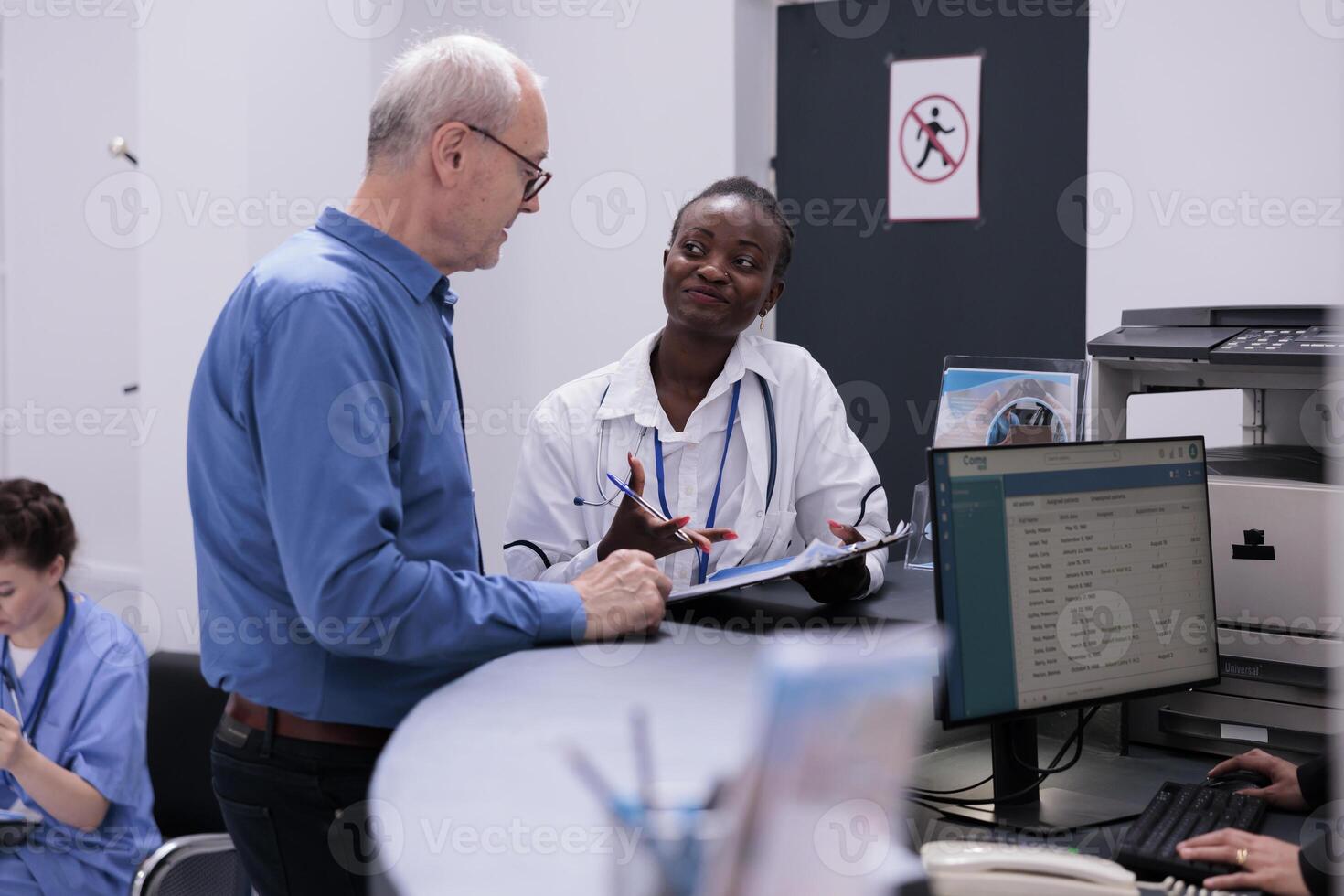 Young adult doctor putting elderly patient to sign medical documents while discussing medication treatment during checkup visit consultation in hospital waiting area. Health care support and concept photo