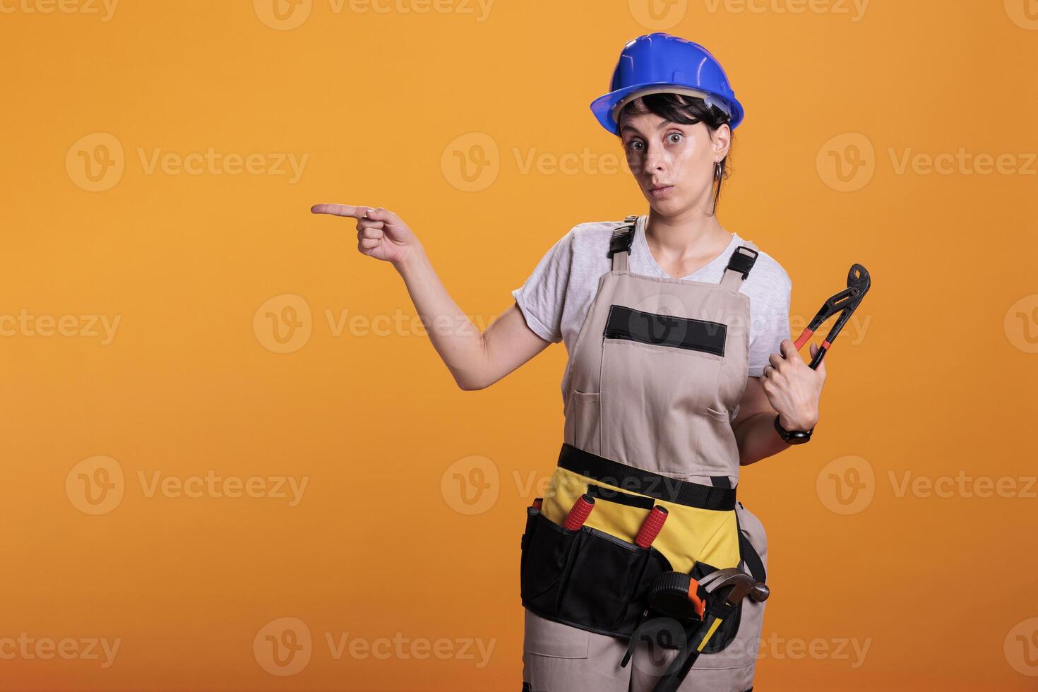 Woman builder indicating direction on left side of studio, posing with pair of pliers and pointing sideways. Female construction worker looking at camera and showing sides, yellow background. photo
