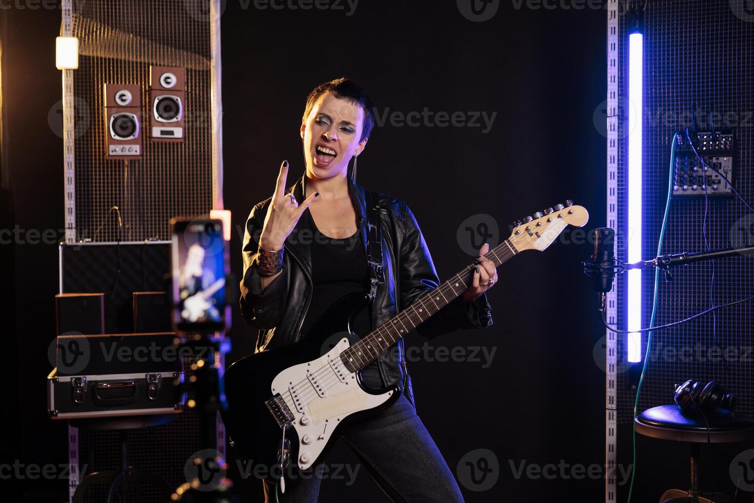 Smiling musician doing rock sign while playing at electric guitar in front of smartphone recording heavy metal song, preparing new rock album. Rebel guitarist performing grunge music in sound studio photo