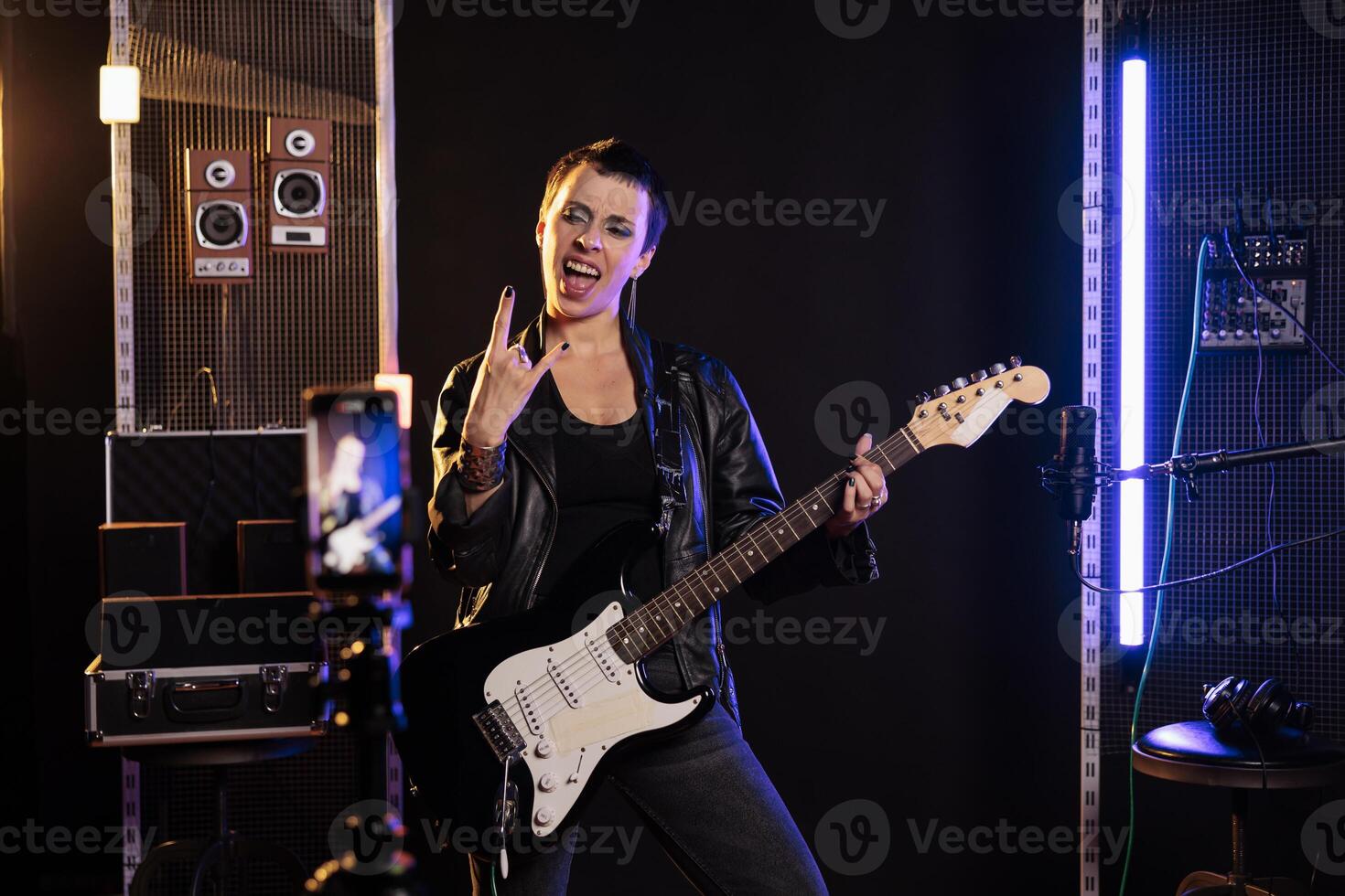 Smiling musician doing rock sign while playing at electric guitar in front of smartphone recording heavy metal song, preparing new rock album. Rebel guitarist performing grunge music in sound studio photo