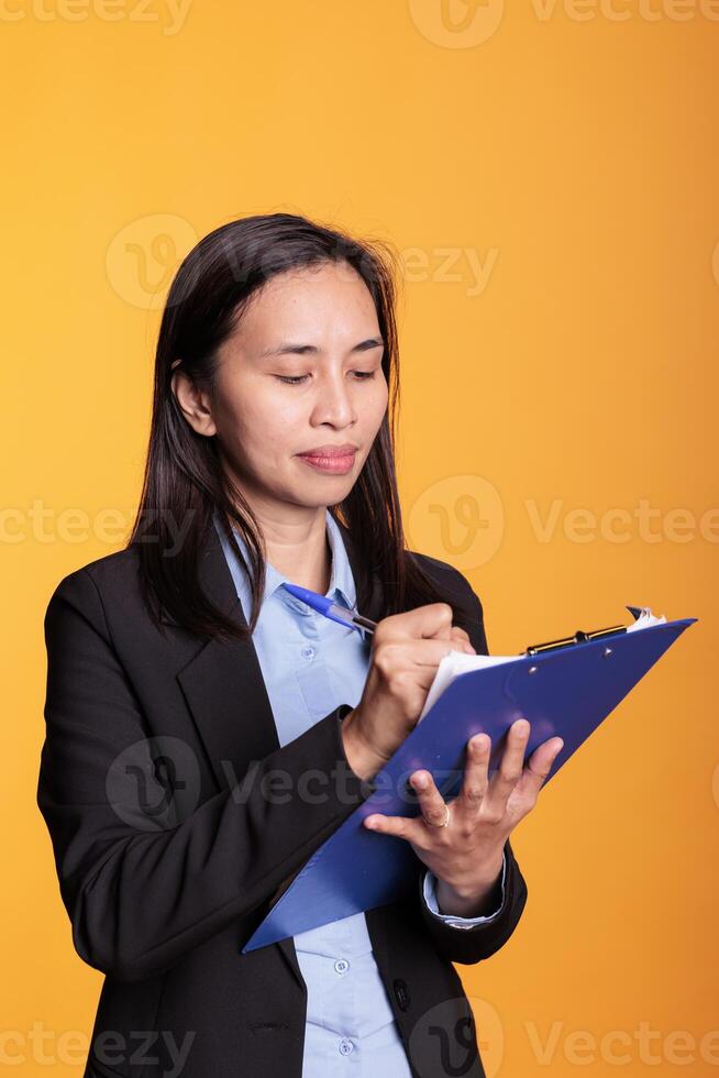 Cheerful woman writing business report on clipboard, filling questionnaire in studio. Filipino attractive adult using papers and pen to create creative project, standing over yellow background photo