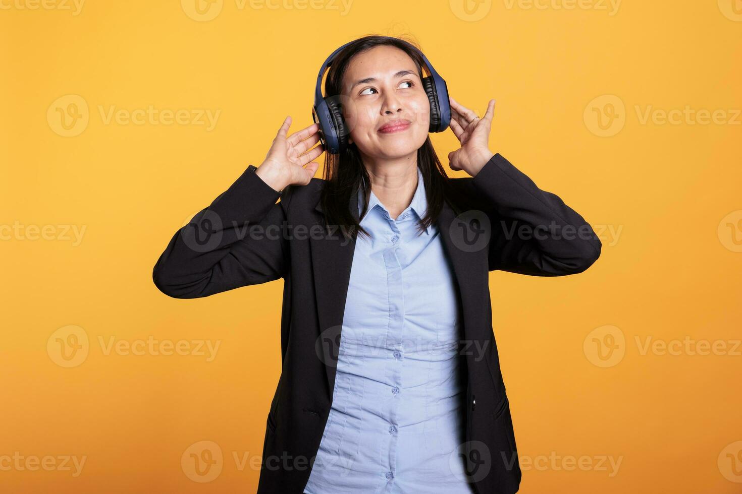 Filipino joyful woman wearing headphones listening music, having fun during break in studio over yellow background. Smiling model enjoying online podcast using headset. Entertainment concept photo