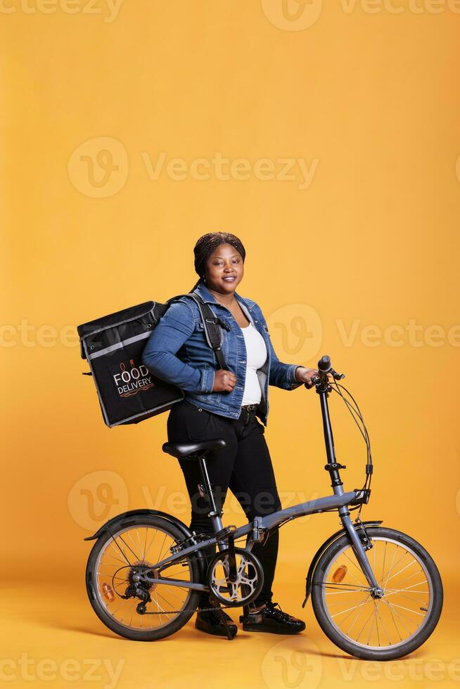 africano americano restaurante mensajero en pie junto a bicicleta participación térmico mochila mientras entregando tomar lejos comida a cliente durante almuerzo tiempo. pizzería empleado que lleva eliminar comida foto