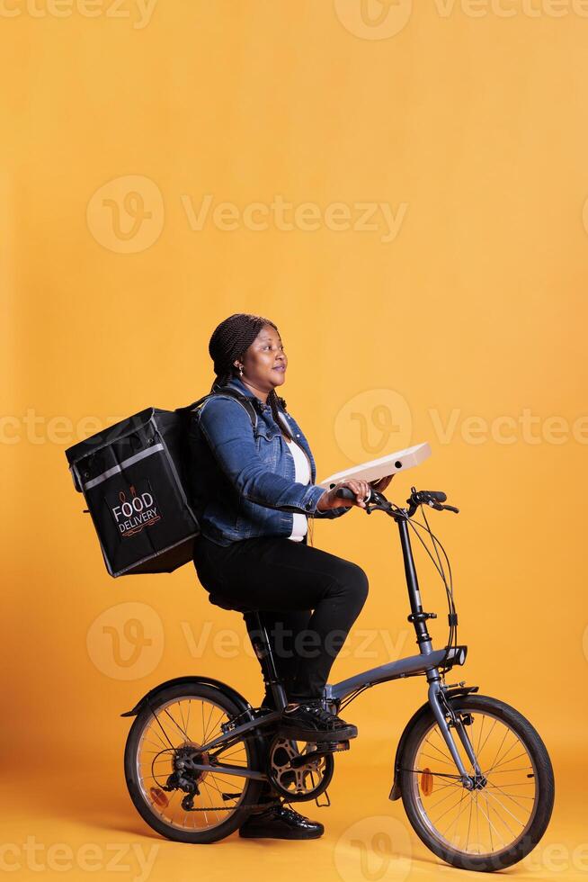 lleno cuerpo de pizzería mensajero participación Pizza cartulina mientras montando bicicleta entregando para llevar orden a cliente durante almuerzo tiempo. restaurante trabajador en pie en estudio con amarillo antecedentes foto