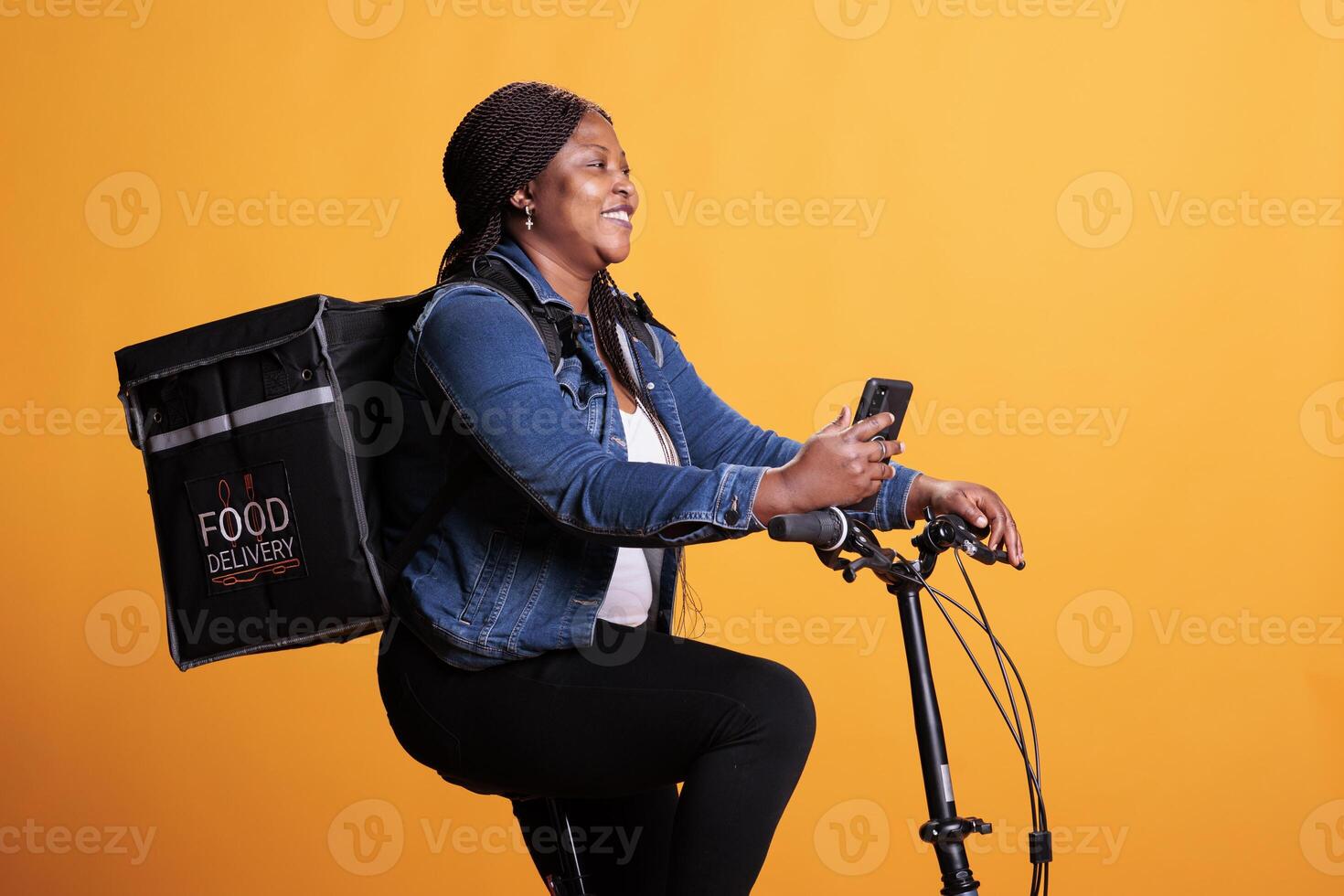 Pizza delivery worker carrying thermal backpack while delivering takeaway food order to customer, holding phone to check address on fast food app. Courier standing over yellow background in studio photo