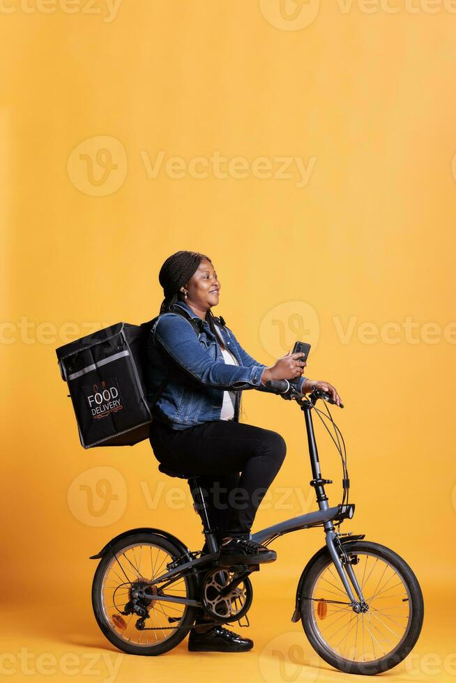 Pizzeria courier riding bike while delivering takeaway food meal to customer, checking adreess on phone takeout app, standing over yellow background. Take out service and concept photo