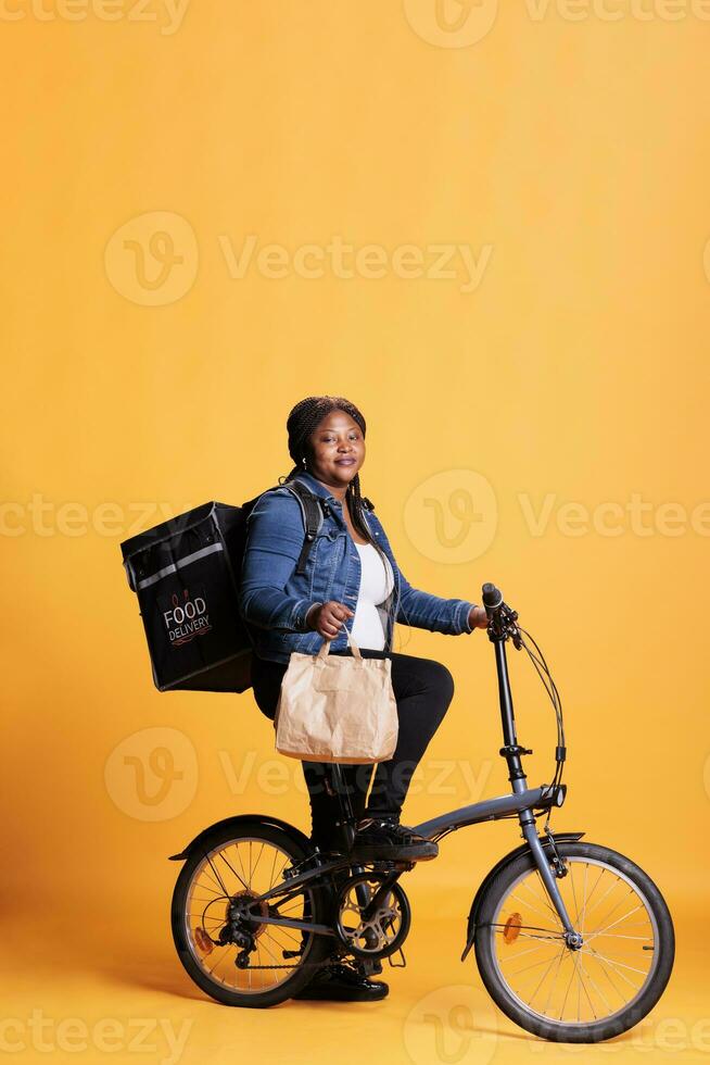 Pizzeria employee holding paper bag with fast food order while delivering lunch to client using bike as transportation. Restaurant courier carrying takeout thermal backpack, takeaway service concept photo