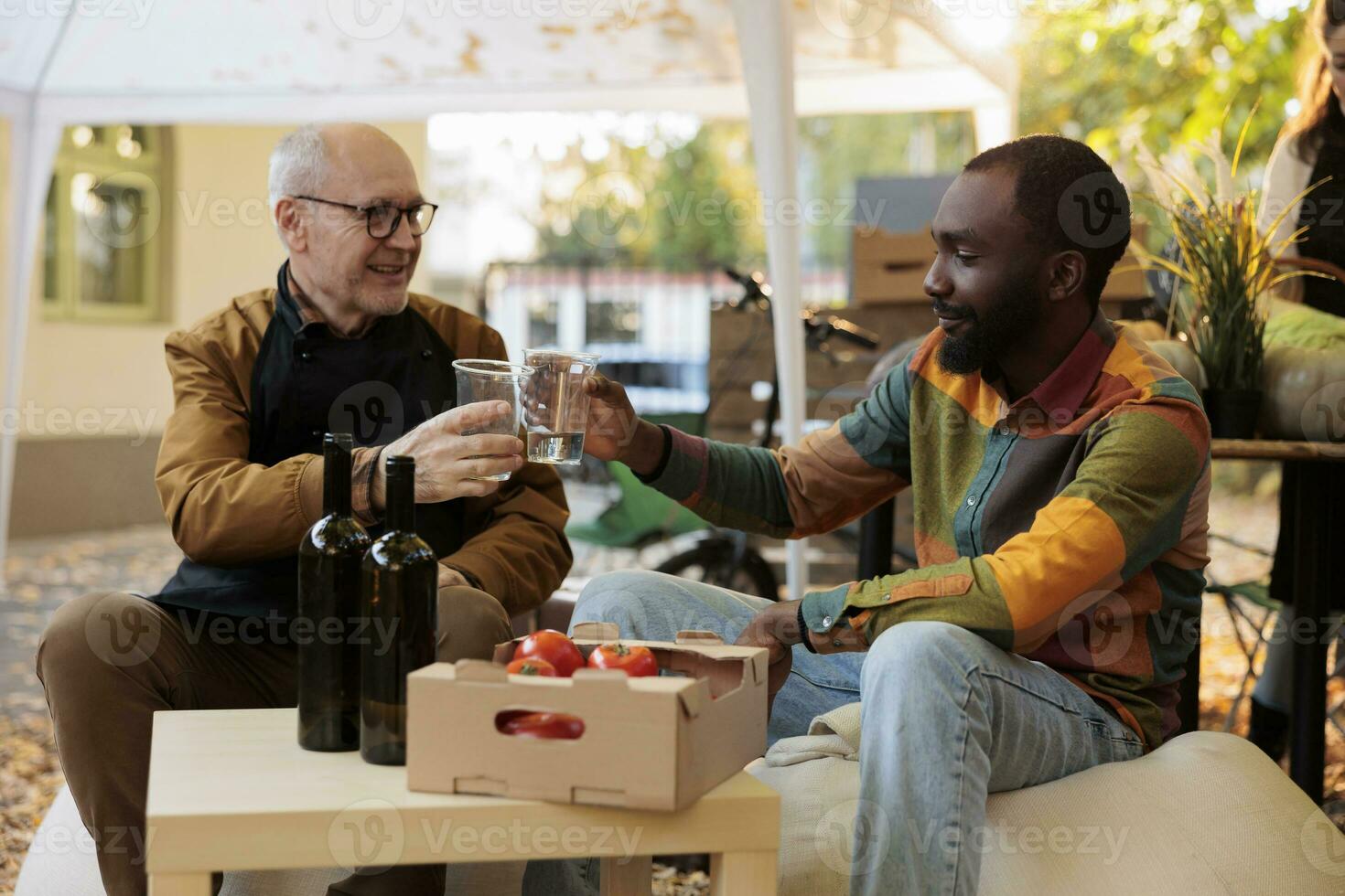 Diverse men clinking glasses of homemade wine at local food fair, tasting organic wine from bottle at farmers market stand. African american man and elderly person drinking bio product. photo